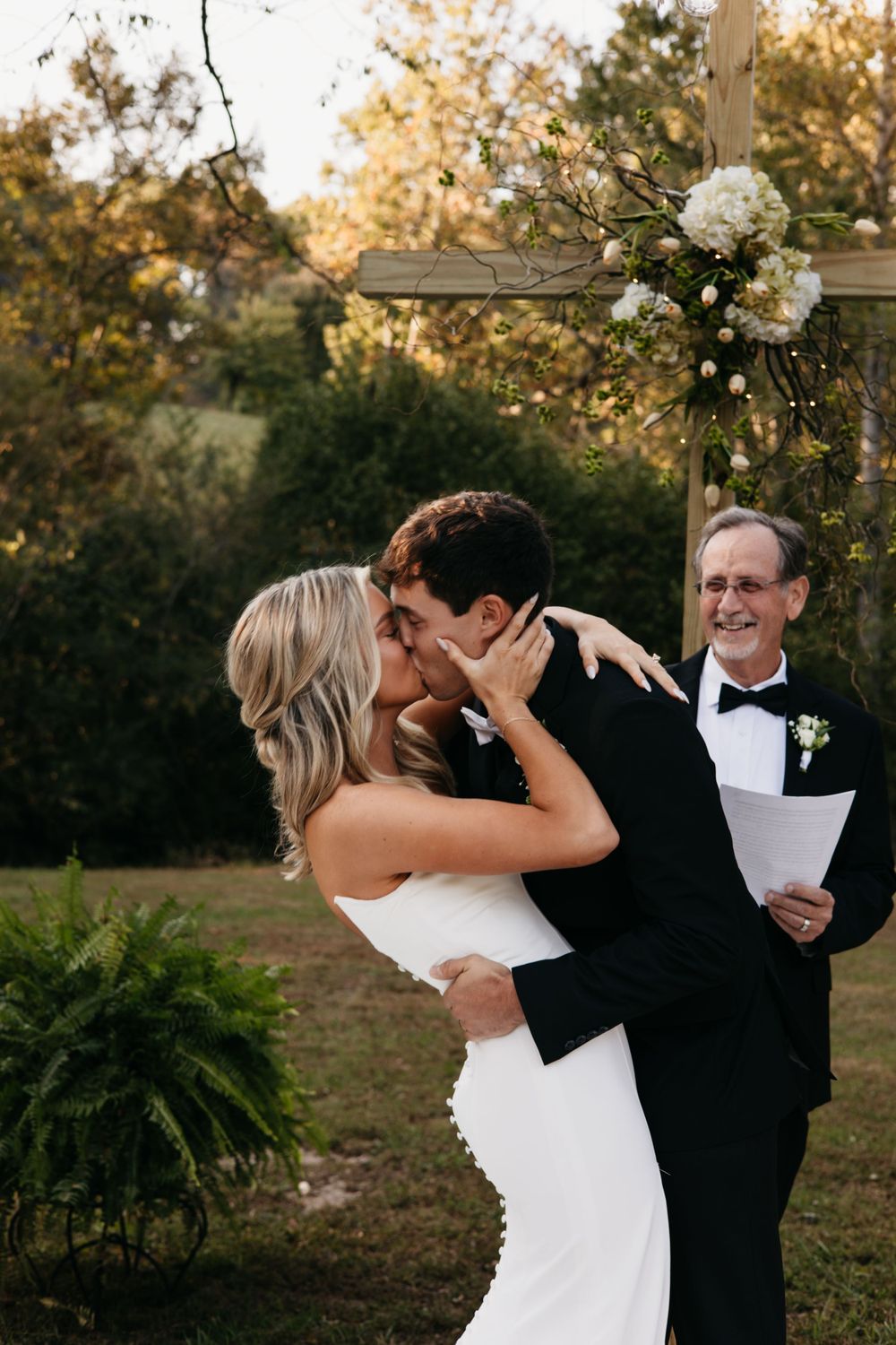 Happy couple shares their first kiss as newlyweds at an outdoor garden ceremony with a smiling officiant.
