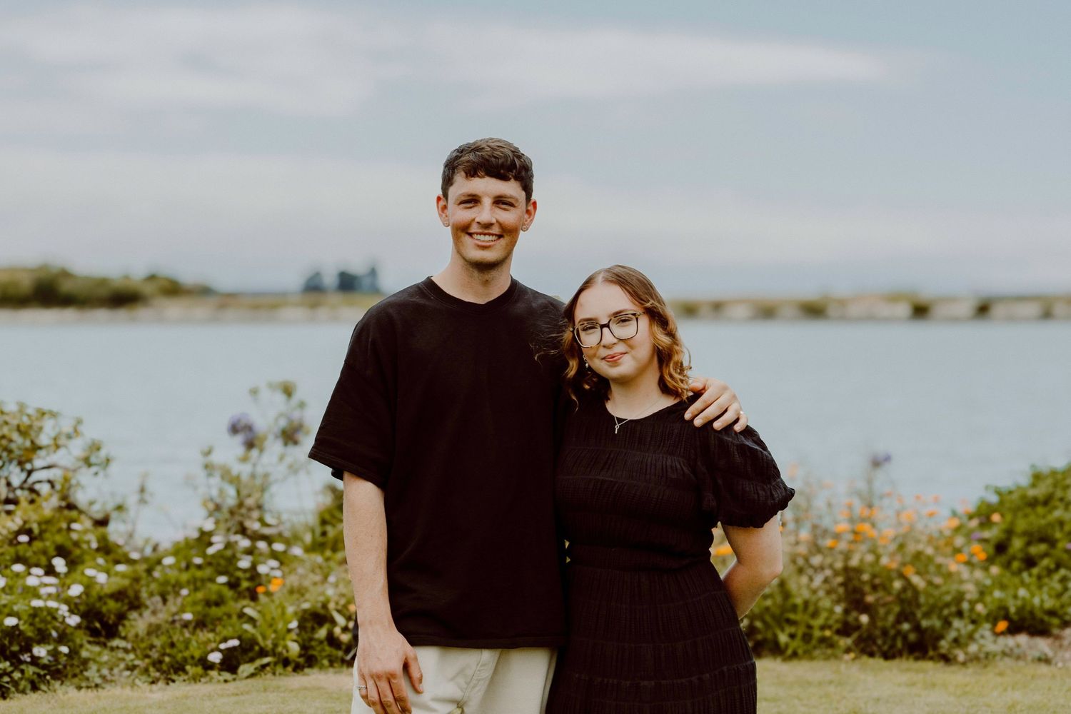 A casual portrait of two friends standing together outdoors by a waterfront.