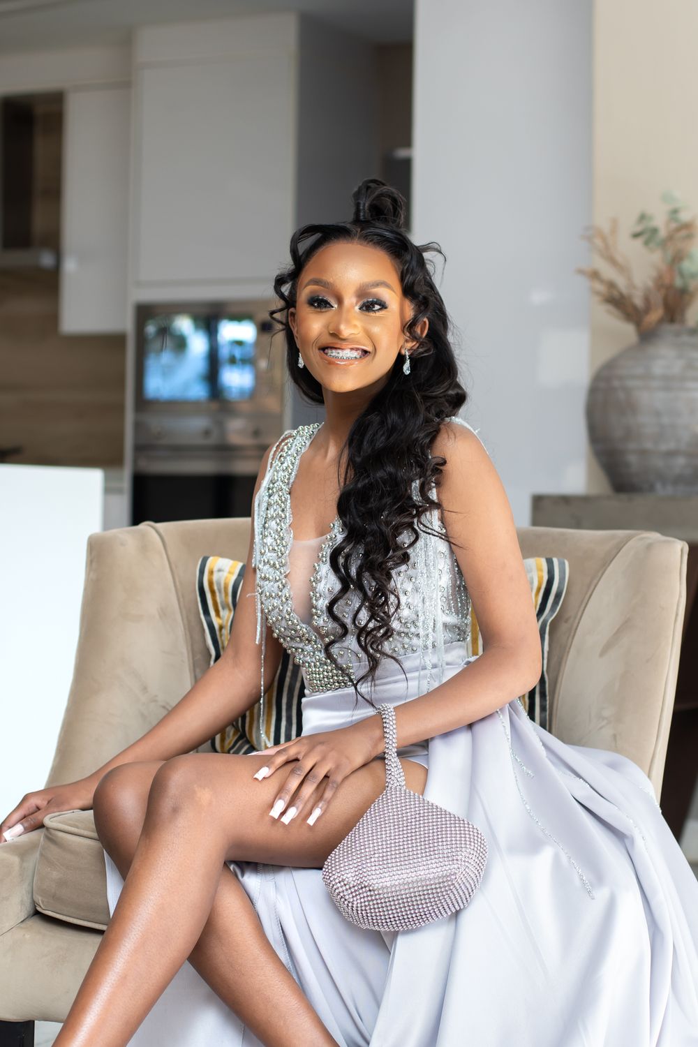 Elegant woman in beaded white gown seated gracefully on upholstered chair.