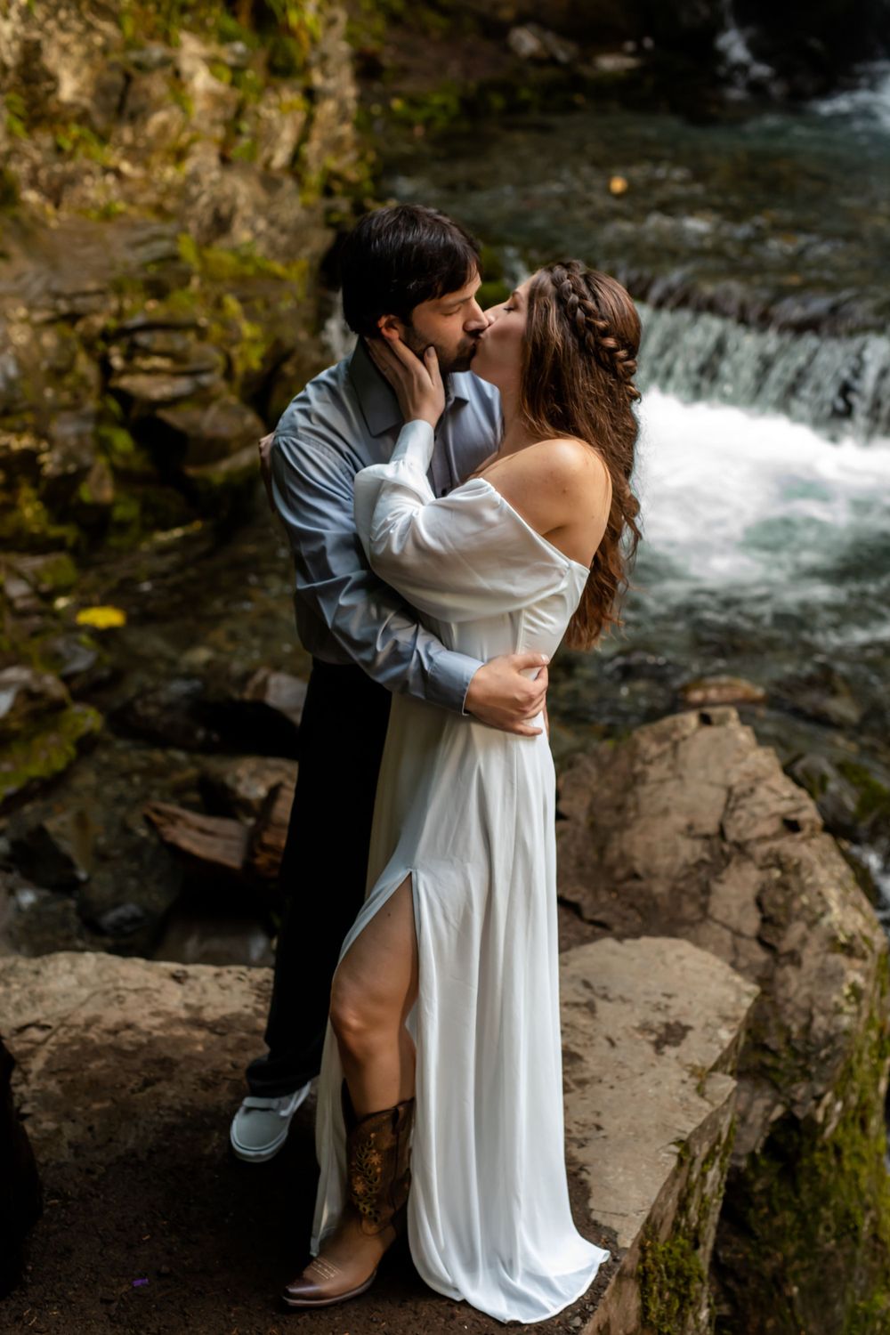 A couple in wedding attire shares intimate moment near cascading waterfall.