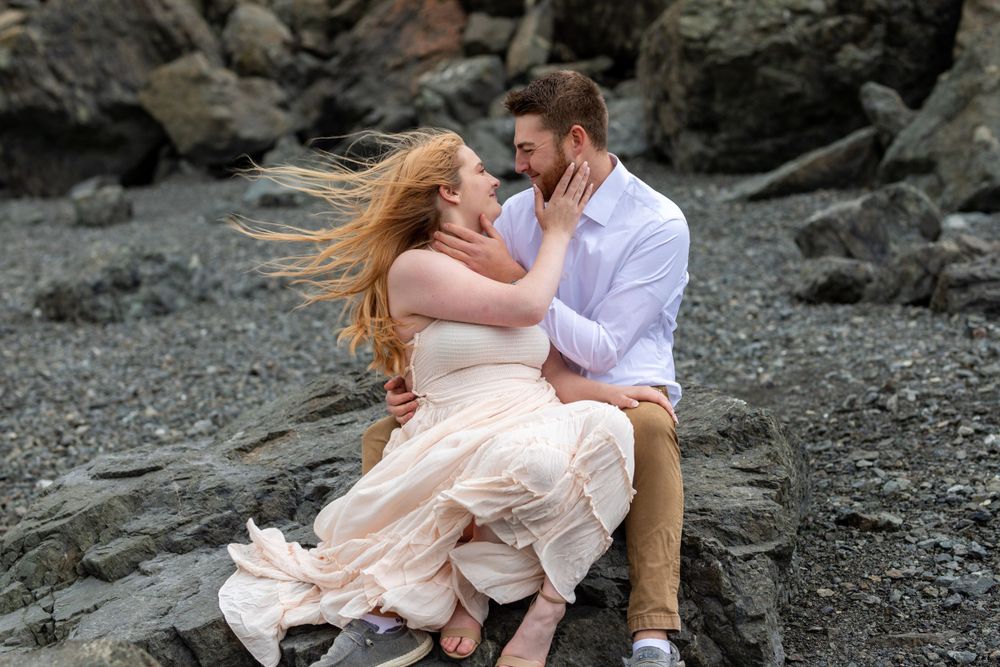 A couple shares a romantic moment on rocky beach with flowing dress and wind-swept hair.