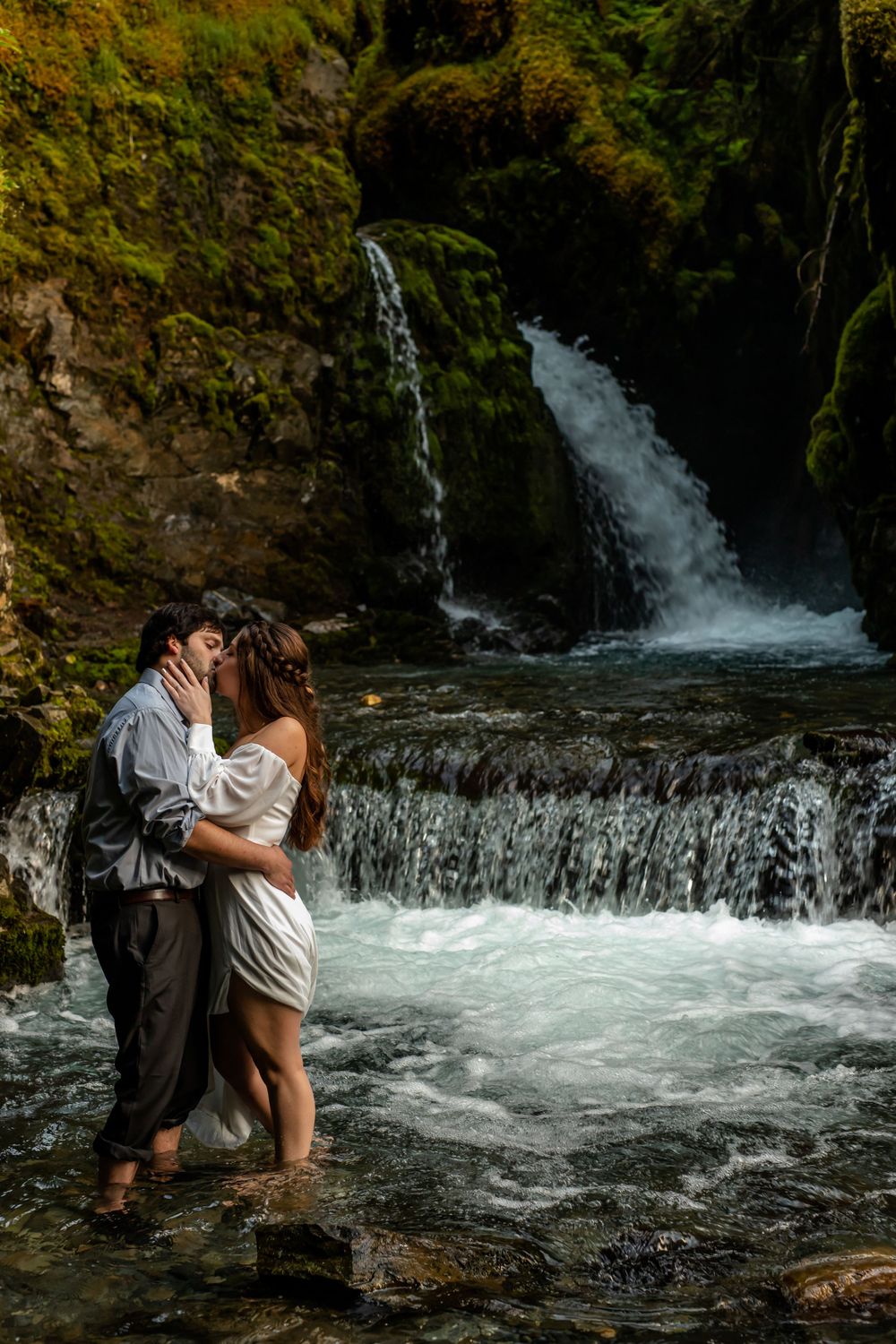 A couple embraces in formal wear while standing in rushing water near a scenic waterfall.