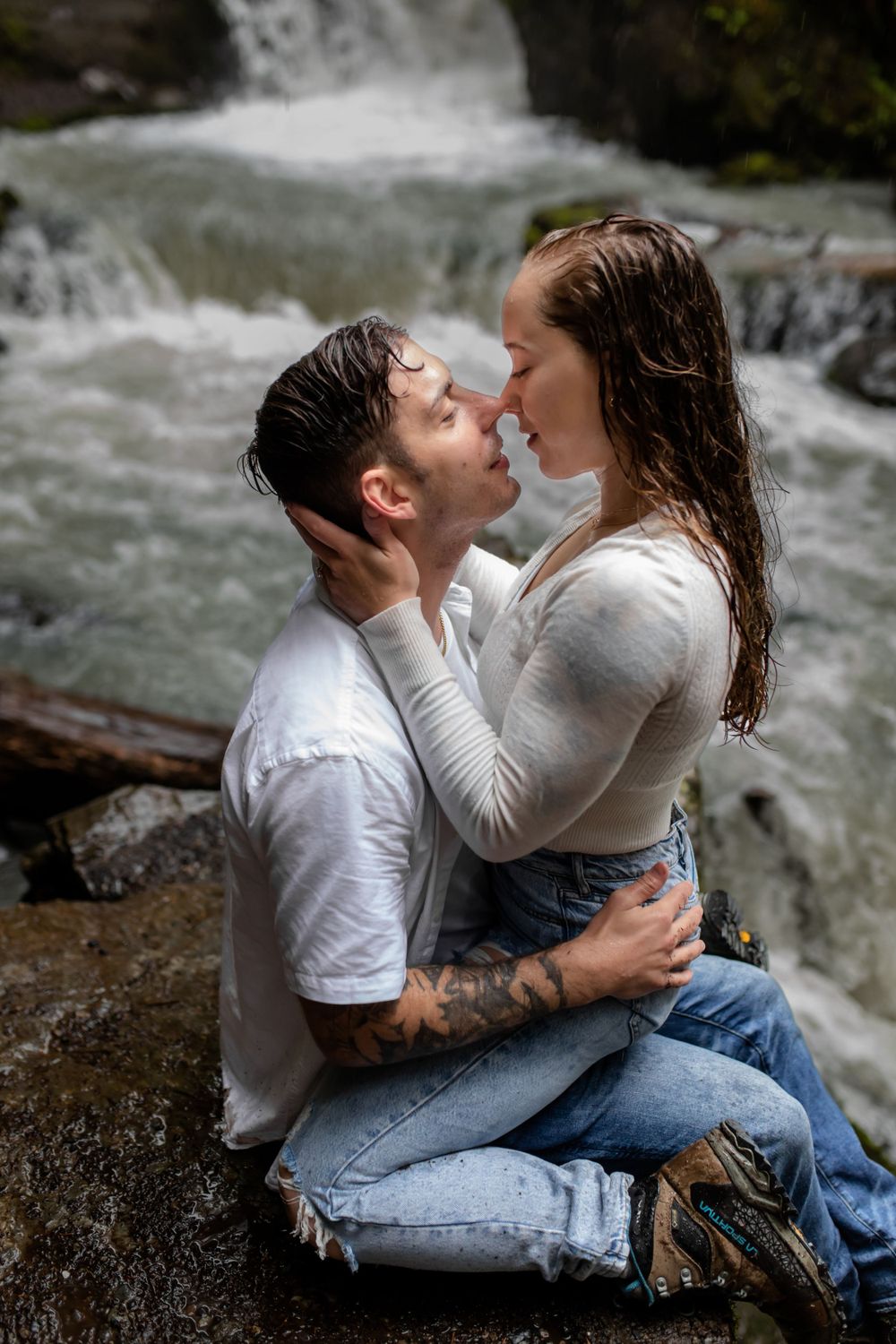 A couple in wet clothes shares intimate moment on rocks by waterfall.