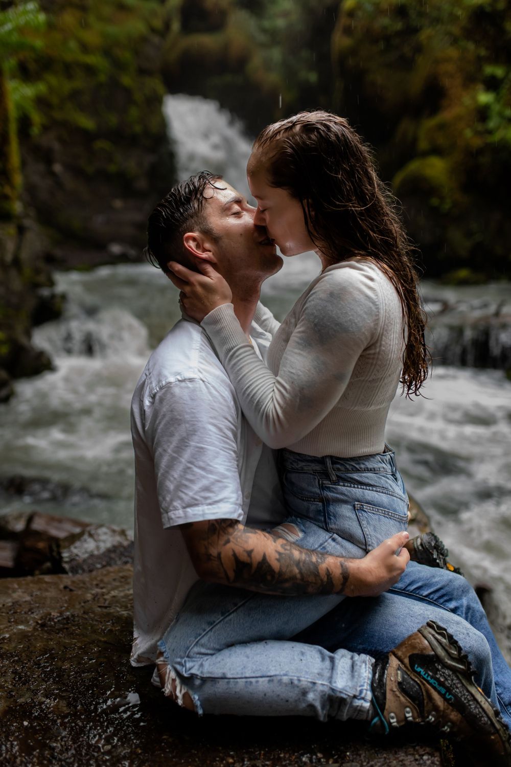 A couple in wet clothes embraces passionately by rushing waterfall.