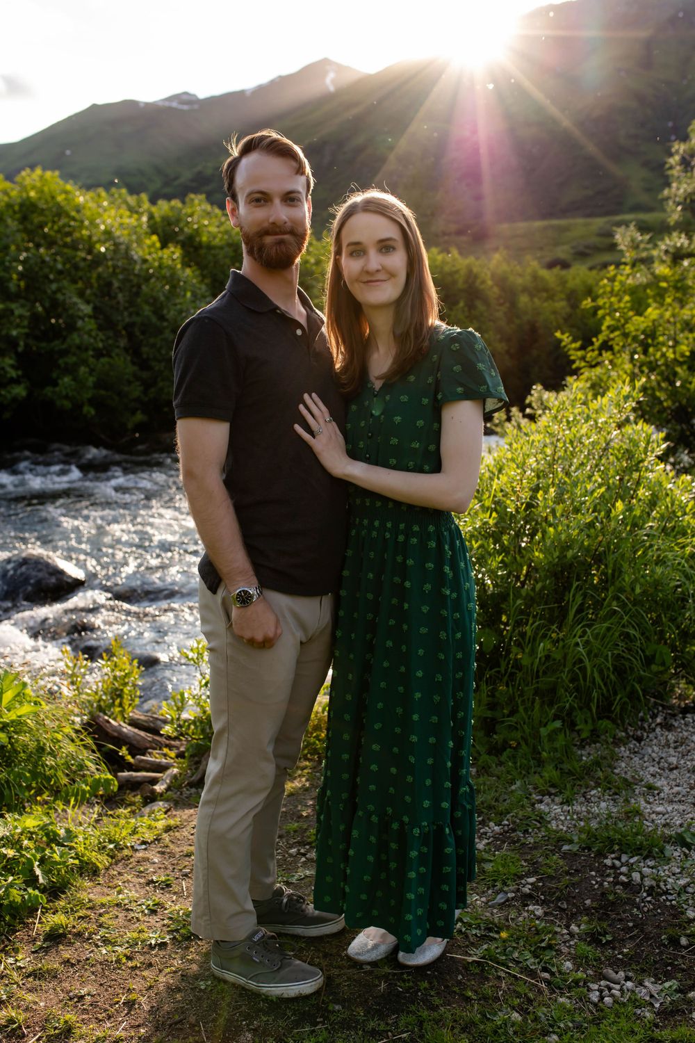 Couple poses together during golden hour near mountain stream with sunburst effect.
