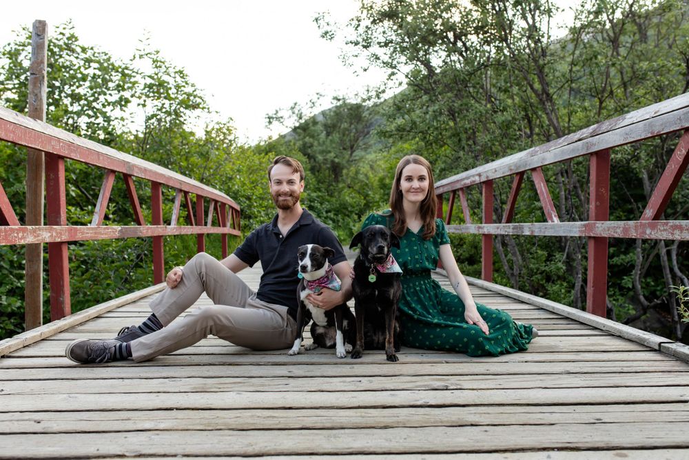 A couple sits on a wooden bridge with their pets on a serene wooded path.