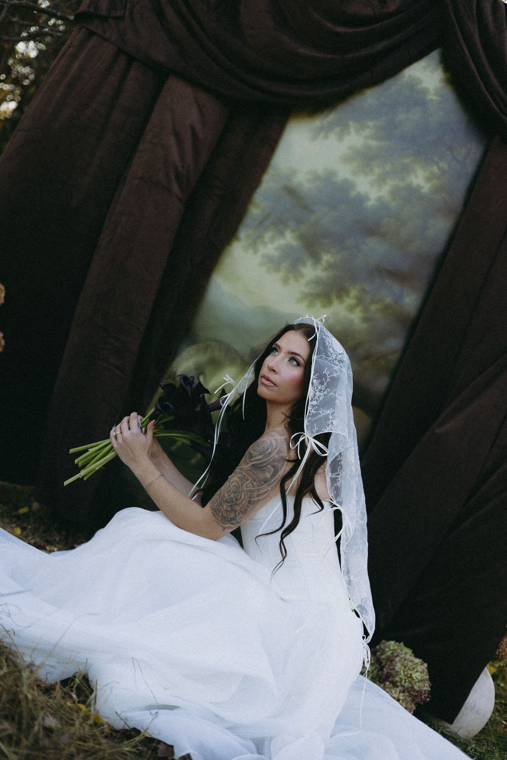 A bride in a white dress and veil sits gracefully holding a bouquet against a dramatic cloudy sky backdrop at her wedding.