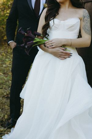Close-up of couple in wedding attire holding dark purple calla lily bouquet, with bride wearing white gown and showing tattoo.