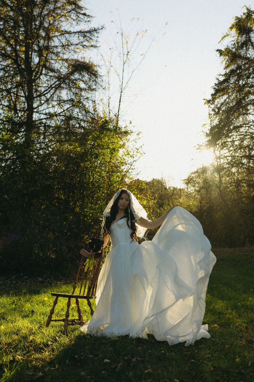 A flowing white wedding dress billows in the sunlight against a backdrop of trees and green grass in a natural setting.
