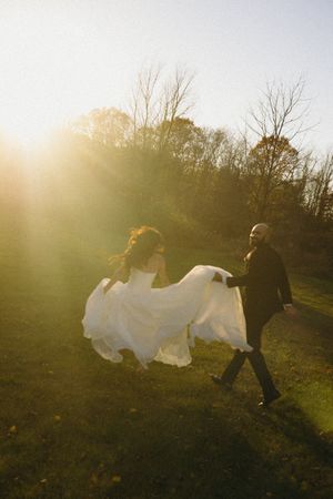 Two figures run through a sunlit field at sunset, with one wearing a flowing white dress that catches the golden light after their wedding.