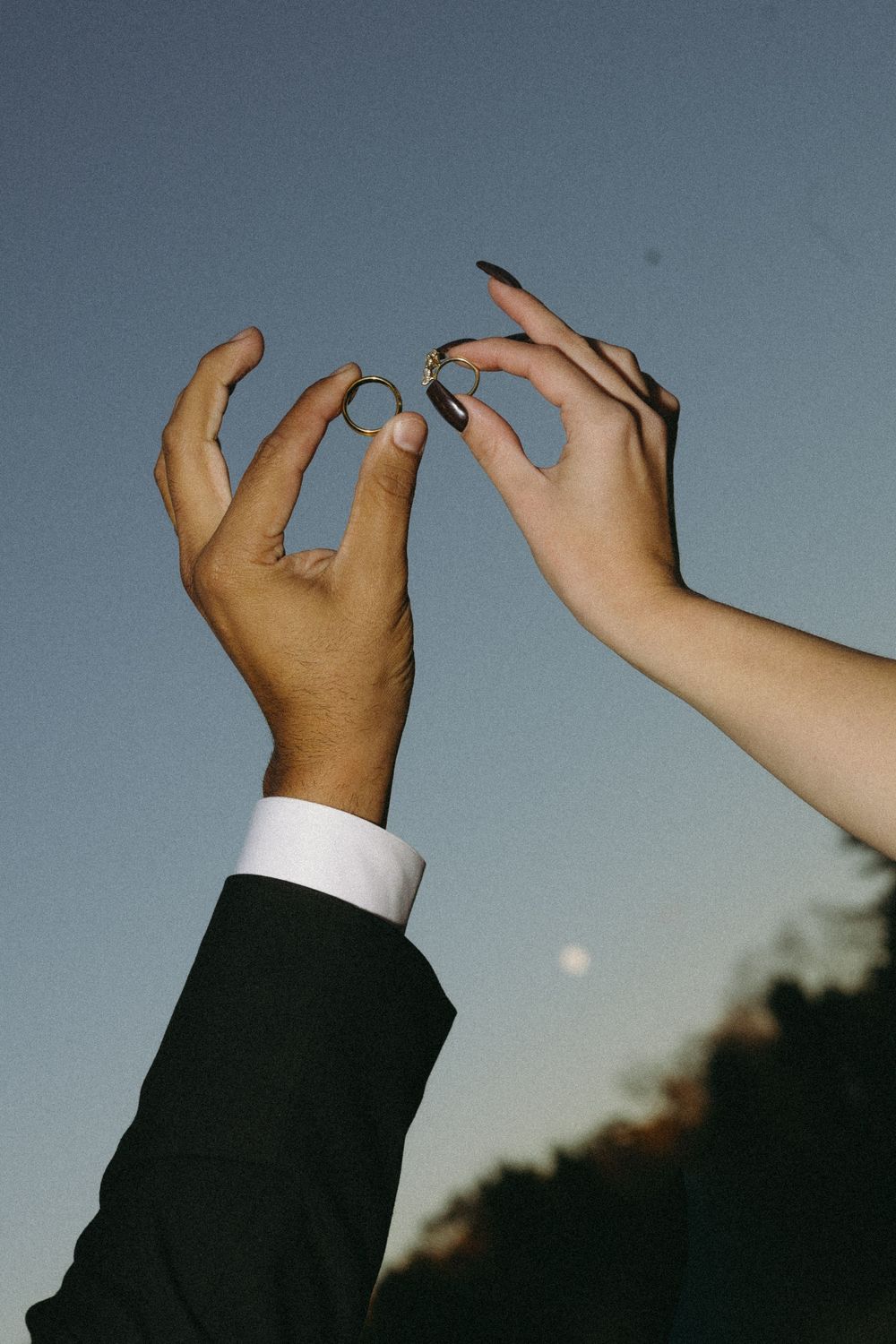 Two raised hands reach toward a dusky sky with the moon visible in the background as they hold up their wedding rings.