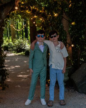 Two figures in sunglasses stand under string lights in a garden setting.
