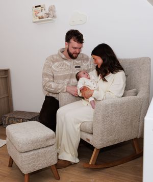 Family sitting together on grey armchair with newborn baby in living room setting.