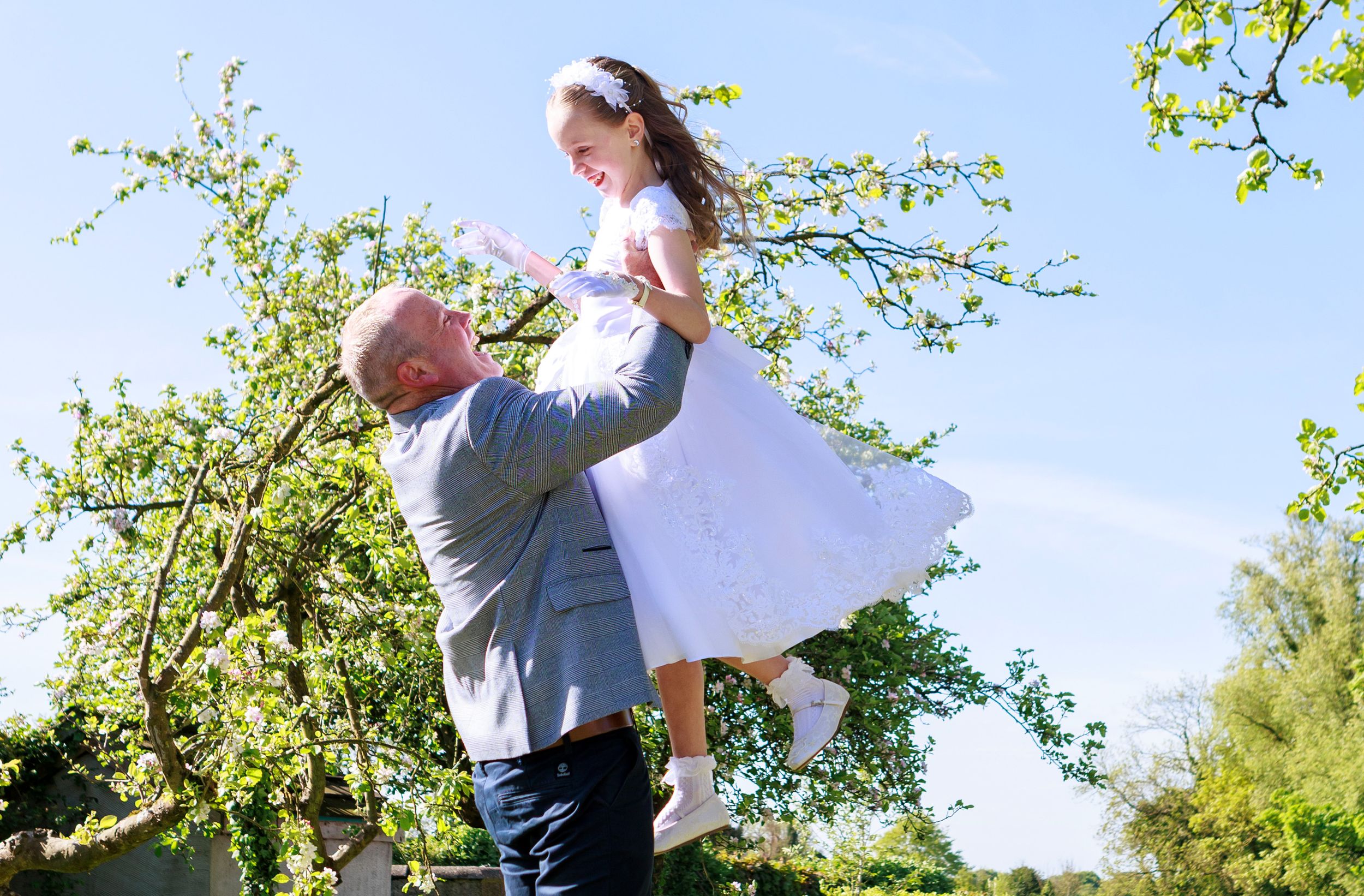 Man lifting young girl in white dress outdoors among green trees on a sunny day.