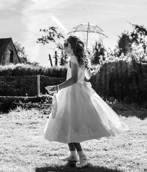 Black and white photo of a young girl in a white dress holding a lace umbrella, twirling in a garden.