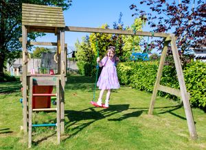 Young girl in pink dress playing on wooden garden swing set with slide on a sunny day.