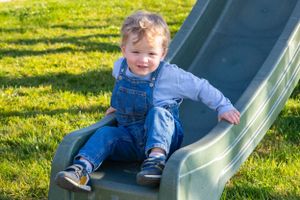 Young child in denim overalls playing on a green plastic slide outdoors on sunny grass.