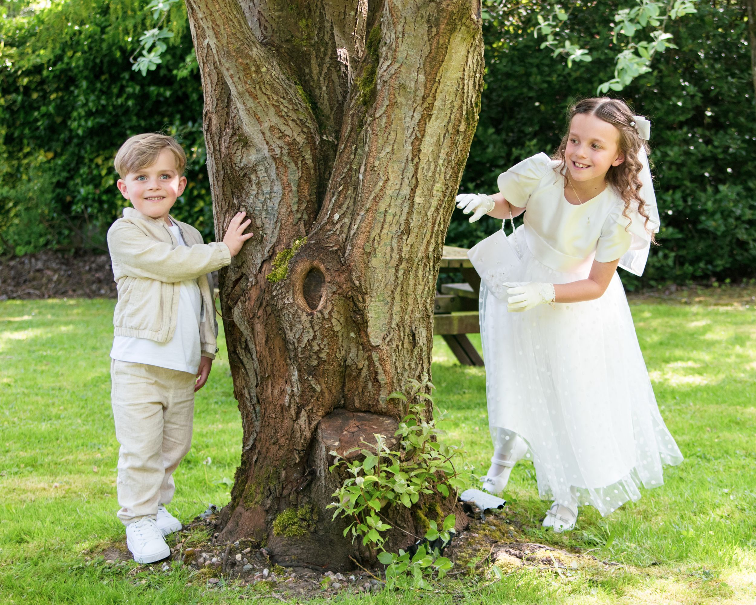 Two children playing peek-a-boo around a large tree trunk in a garden setting.