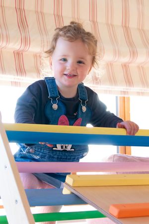 Toddler in overalls smiling and playing at a colorful activity table indoors.