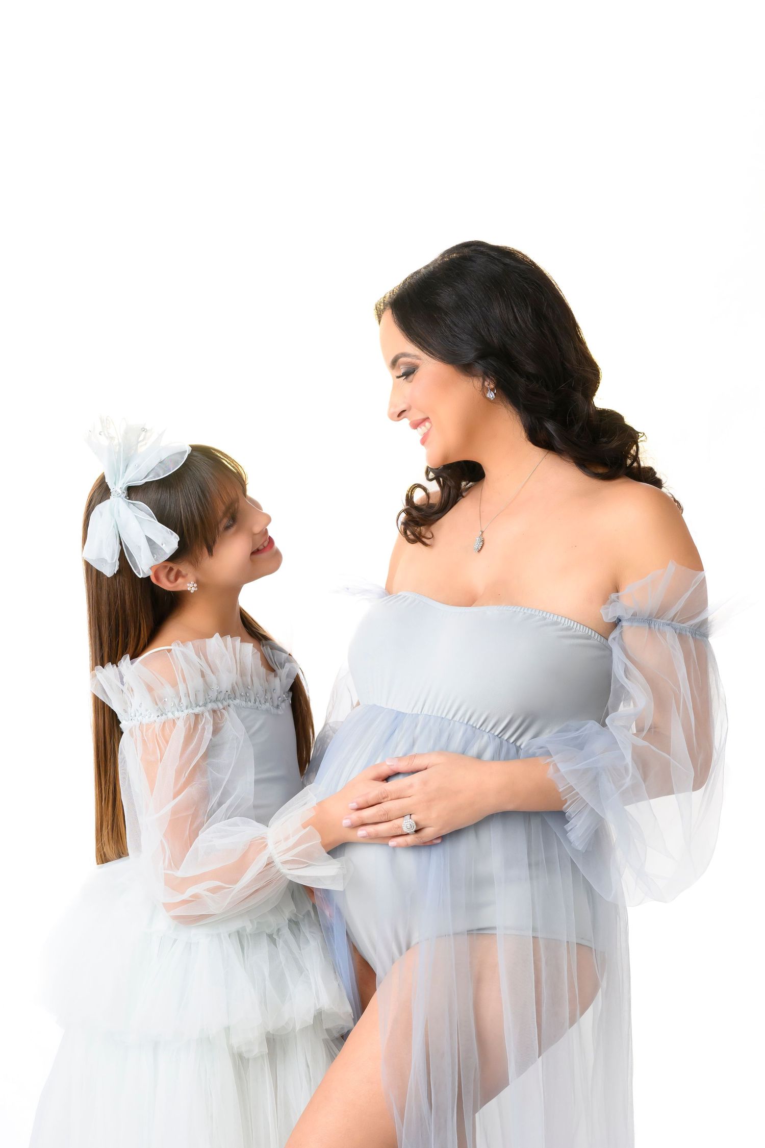 A mother and daughter share a tender moment wearing matching light blue tulle dresses against a white background.