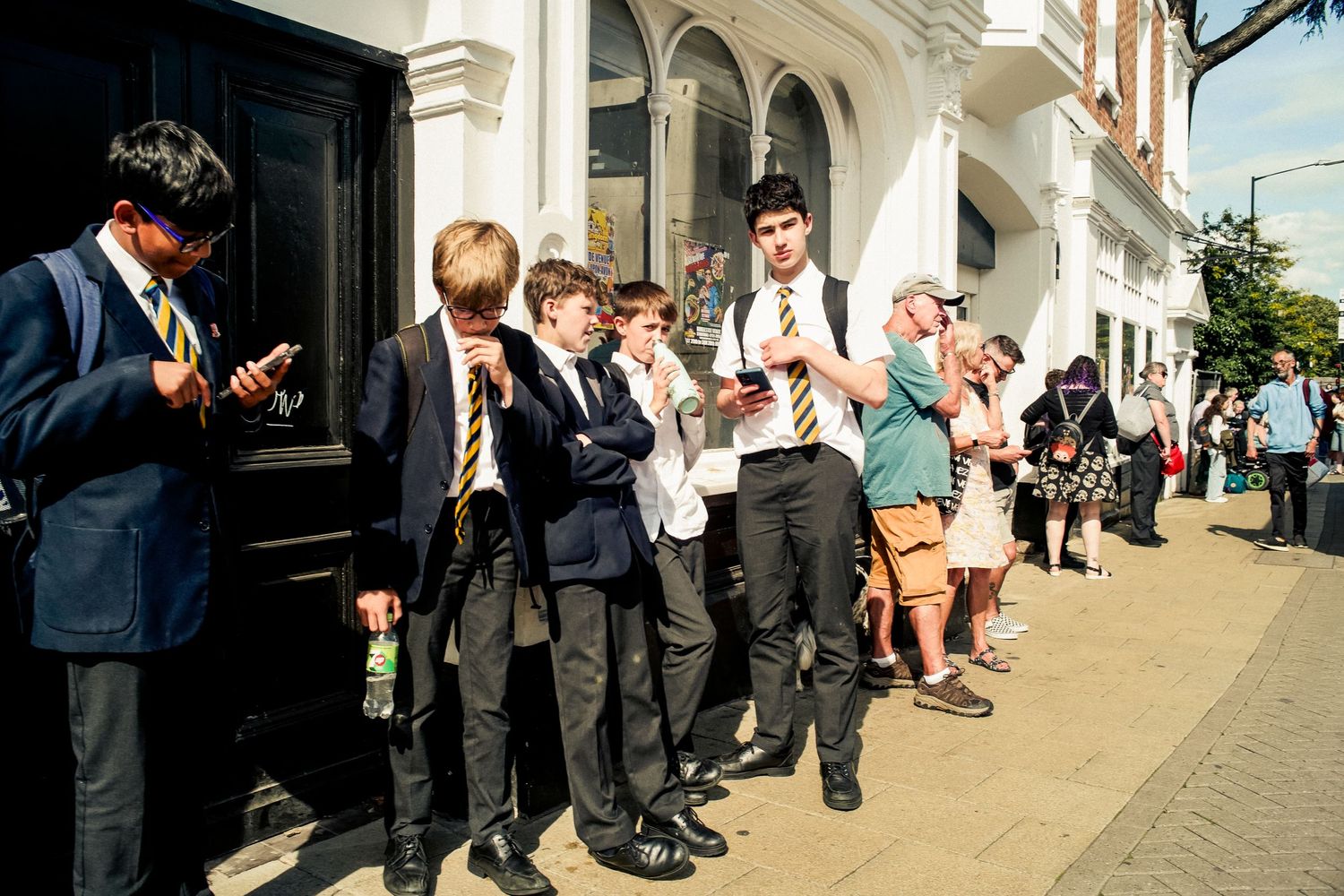 Students in uniforms standing along a building wall checking their mobile phones.