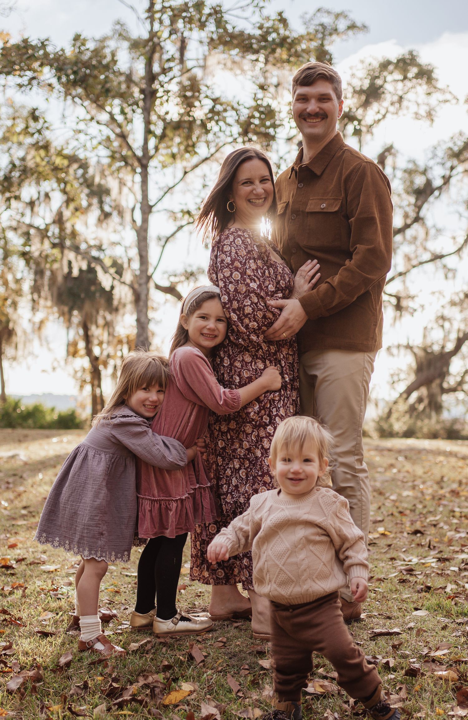A family poses together outdoors on fallen autumn leaves with sunlight filtering through trees in the background.