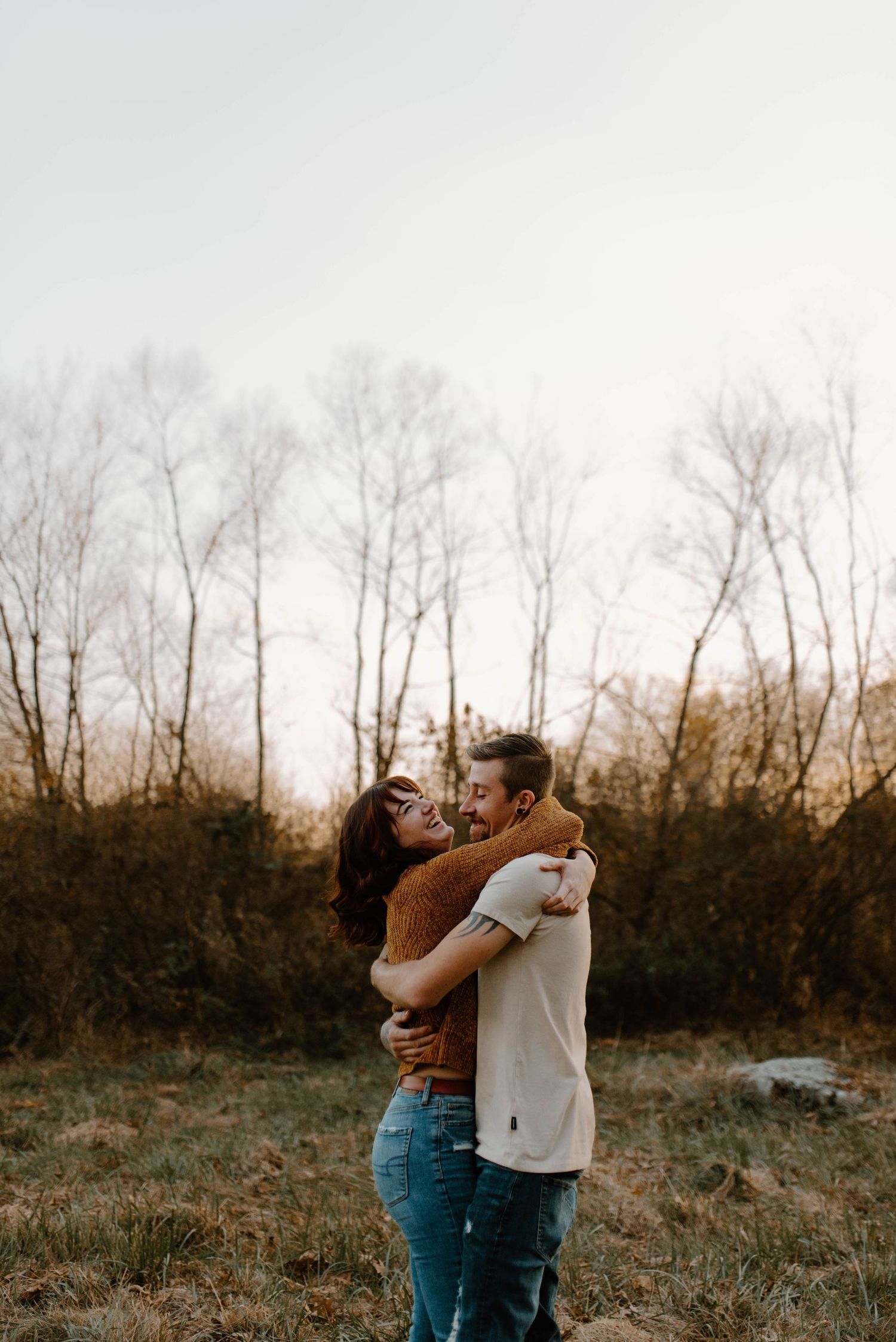 A romantic couple embraces in a field at sunset with bare trees in the background.