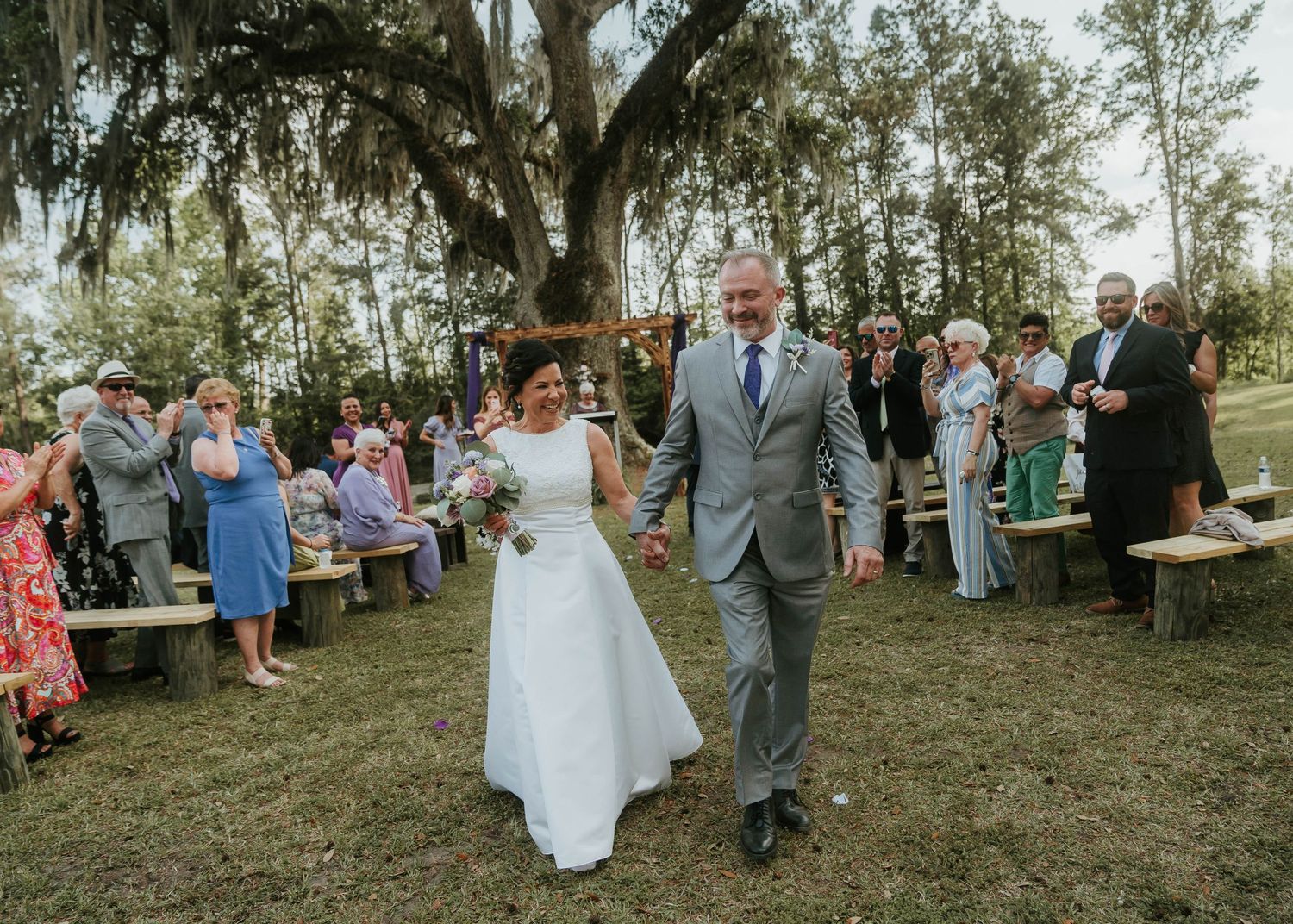 A wedding celebration takes place outdoors under Spanish moss-draped trees with guests seated on wooden benches.