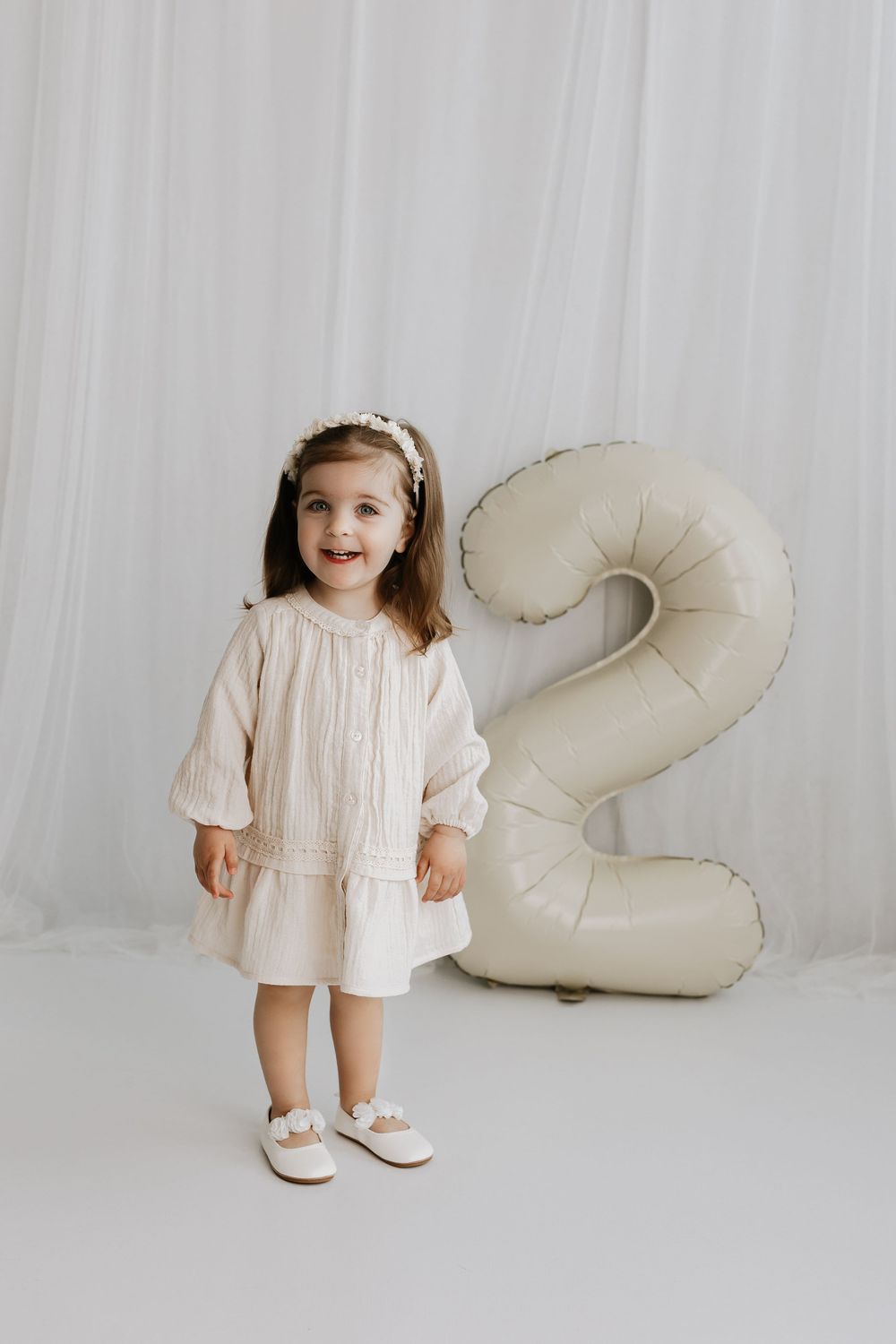 A toddler in a cream-colored dress stands beside a large white inflatable number 2.