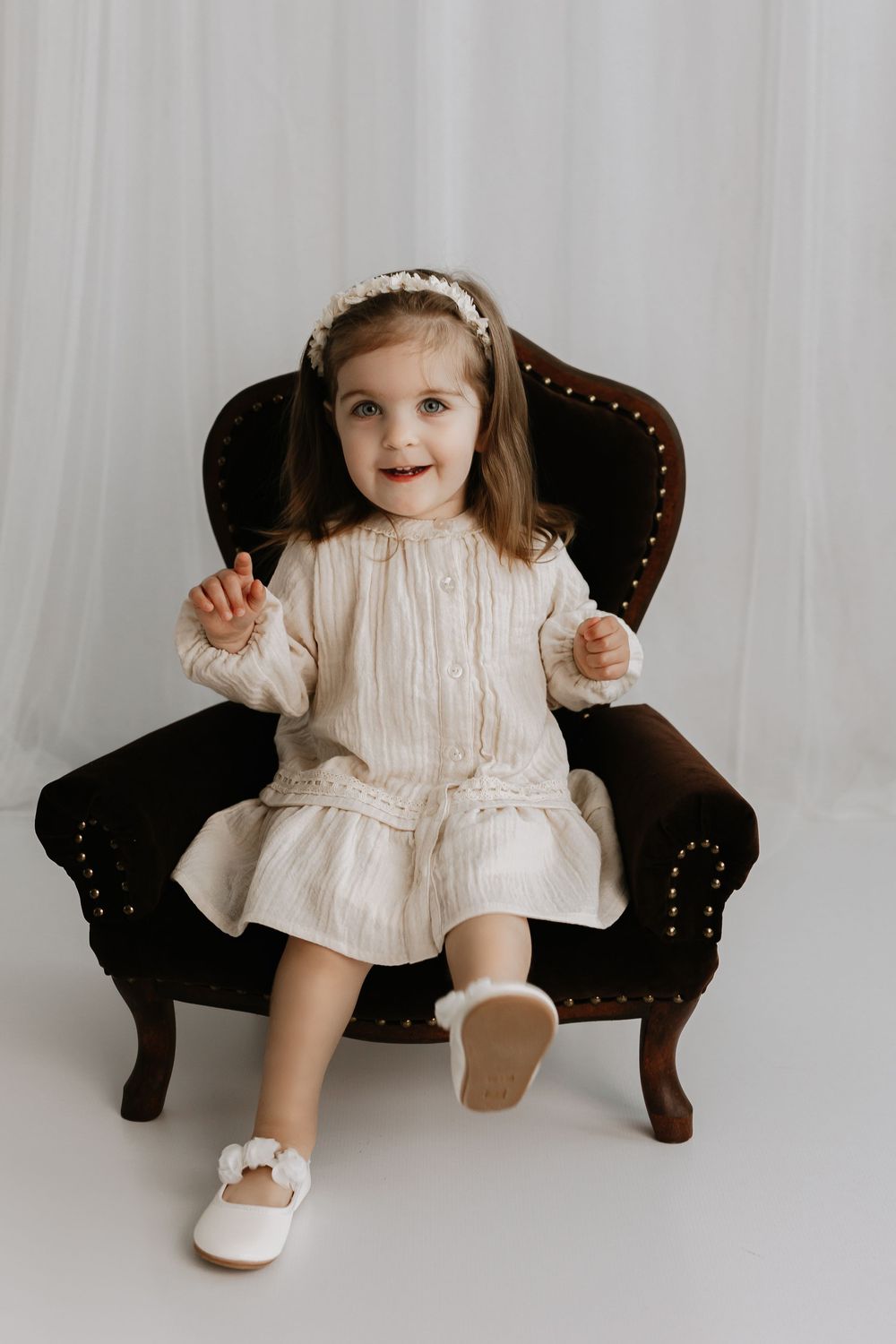 A little one sits in a dark vintage armchair wearing a light-colored dress against a white background.