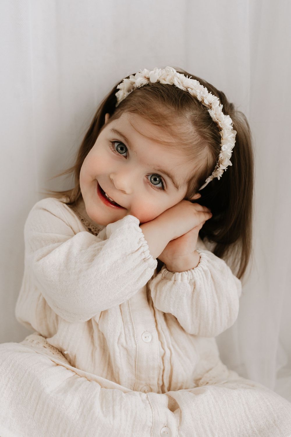 Happy child in cream colored dress with floral headband poses sweetly against light background.