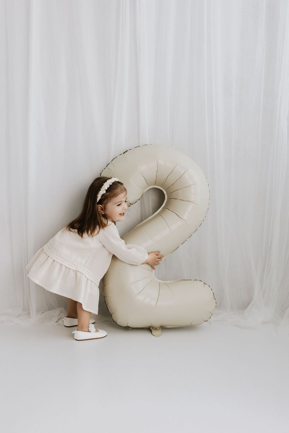 A child in a cream dress plays with a large inflatable number 2 against a white backdrop.