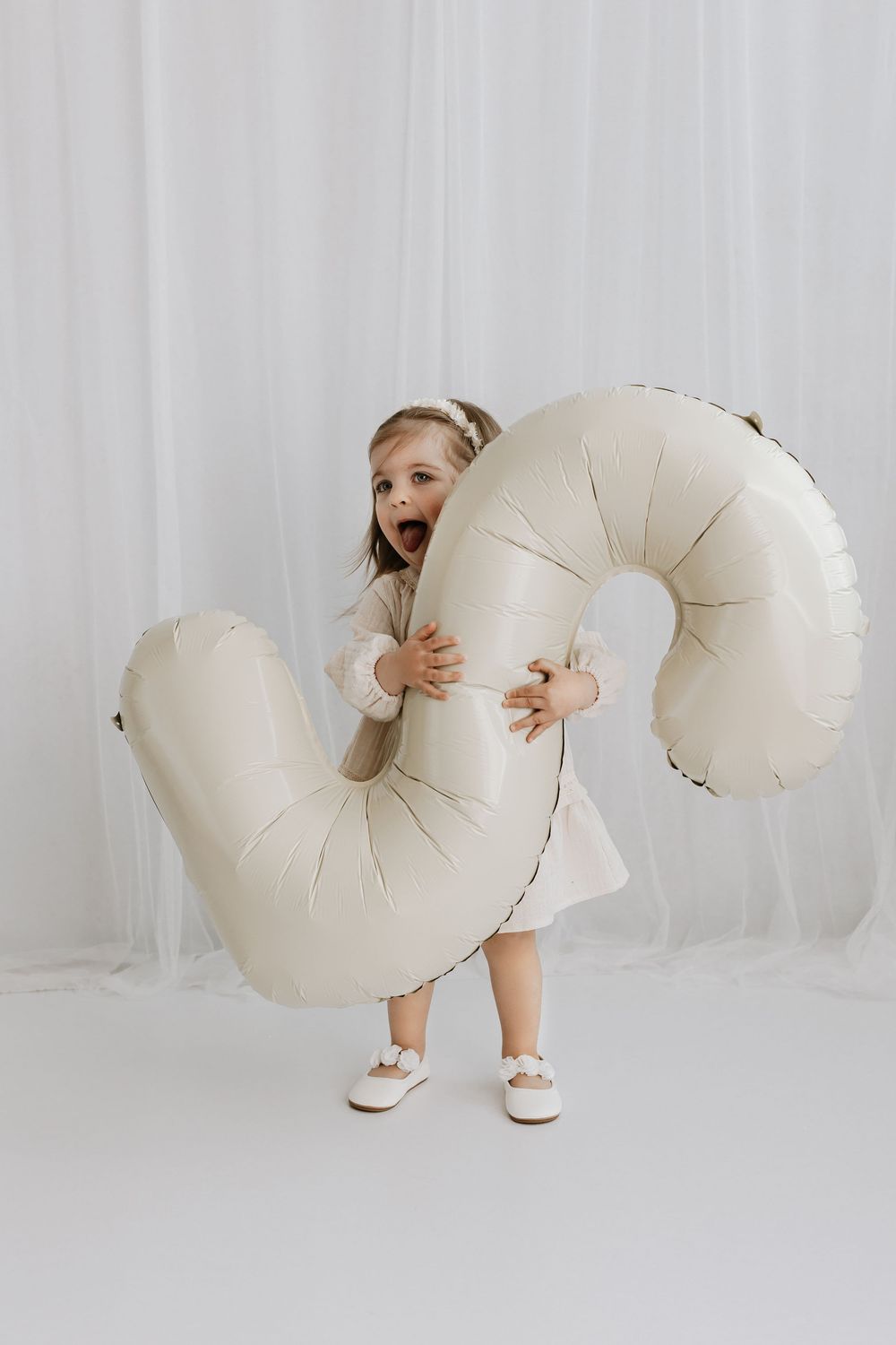 Child in white dress plays with large cream colored, spiral-shaped inflatable prop against white backdrop.