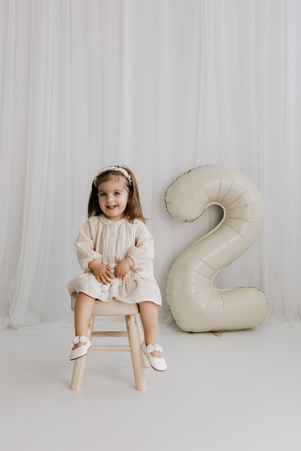 Child in white dress sits on wooden stool next to large number 2 balloon for birthday photo shoot.