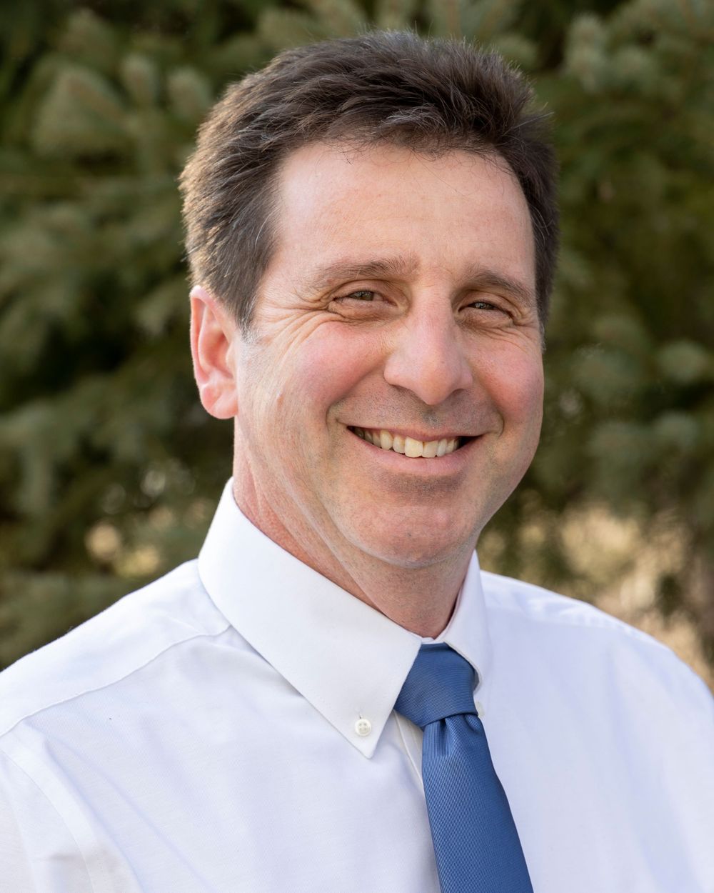 Professional headshot of someone in a white dress shirt and blue tie smiling warmly at the camera in an outdoor setting.