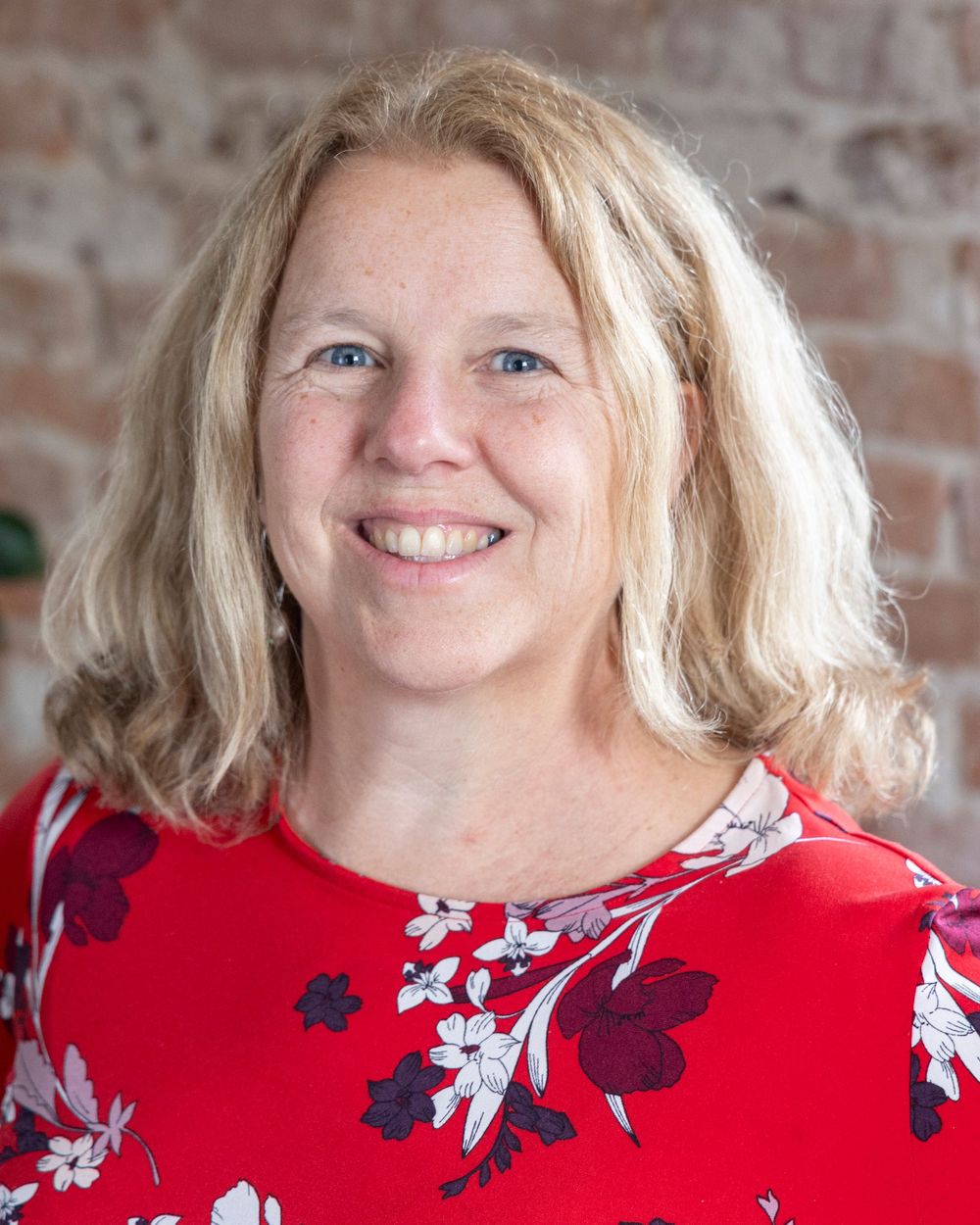 Professional headshot of someone in a red floral top with flowing blonde hair against a textured brick wall background.