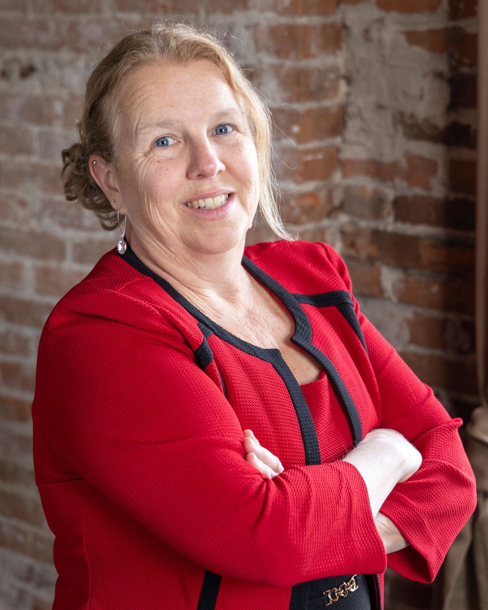 A person wearing a bright red blazer stands with crossed arms against a rustic exposed brick wall background.
