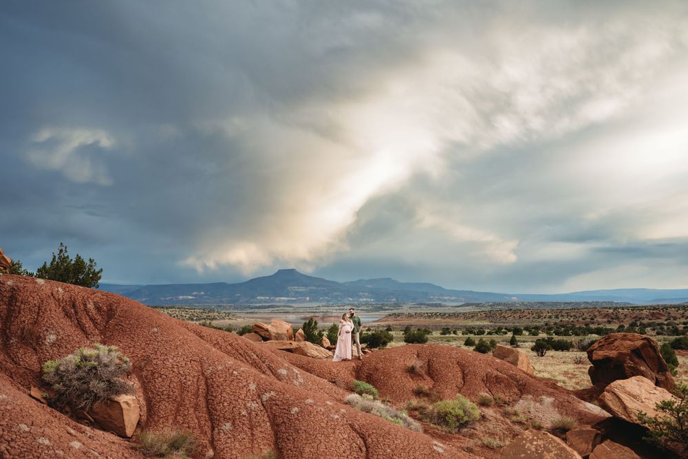 Couple on red desert rocks in New Mexico, with a dramatic cloudy sky and mountain views, captured during a maternity photography session.