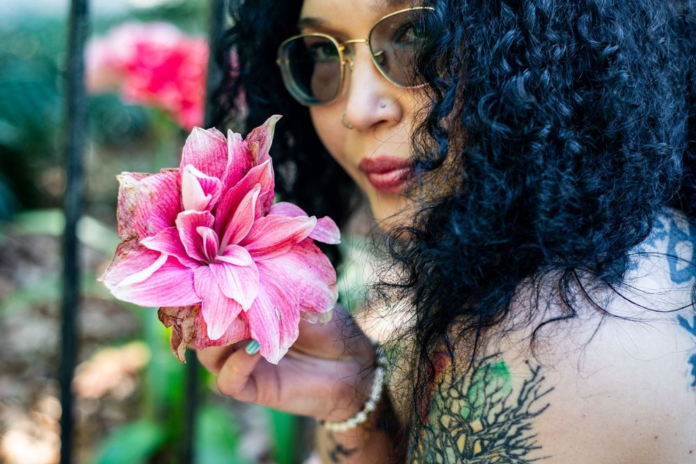 Person with dark curly hair poses near vibrant pink tropical flowers in a lush garden setting.