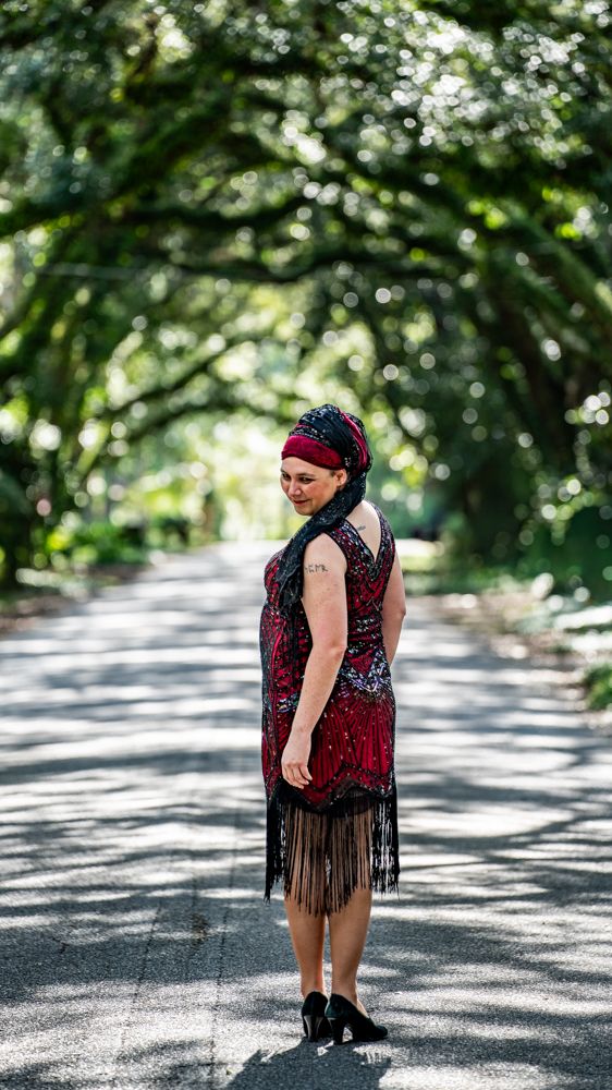 A person wearing a red and black fringed dress stands on a tree-lined path in dappled sunlight.
