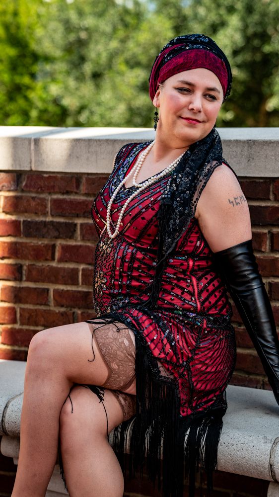 A person poses outdoors wearing a red and black sequined dress with black gloves against a brick wall.