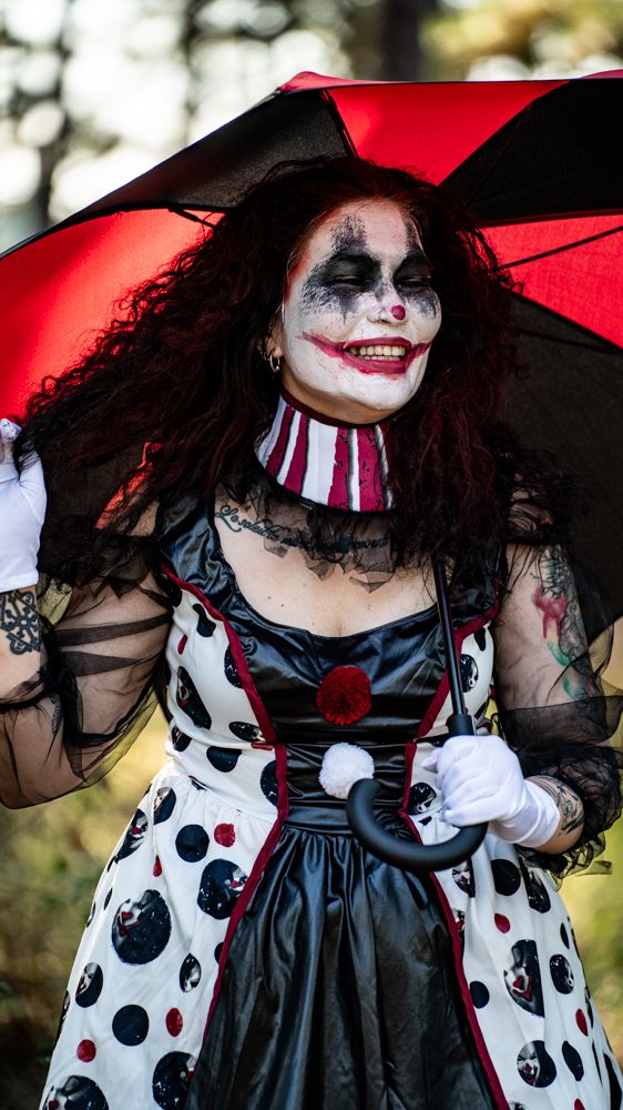A person in dramatic clown makeup and costume with black and white polka dots poses with a red umbrella outdoors.