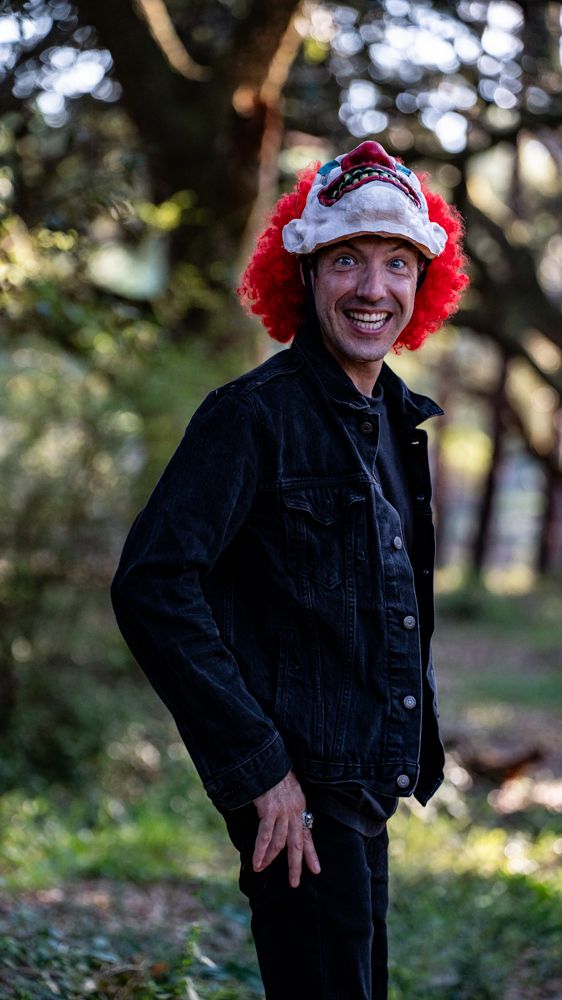 Someone wearing a black denim jacket and festive red clown wig with a Santa hat stands outdoors in a wooded setting.