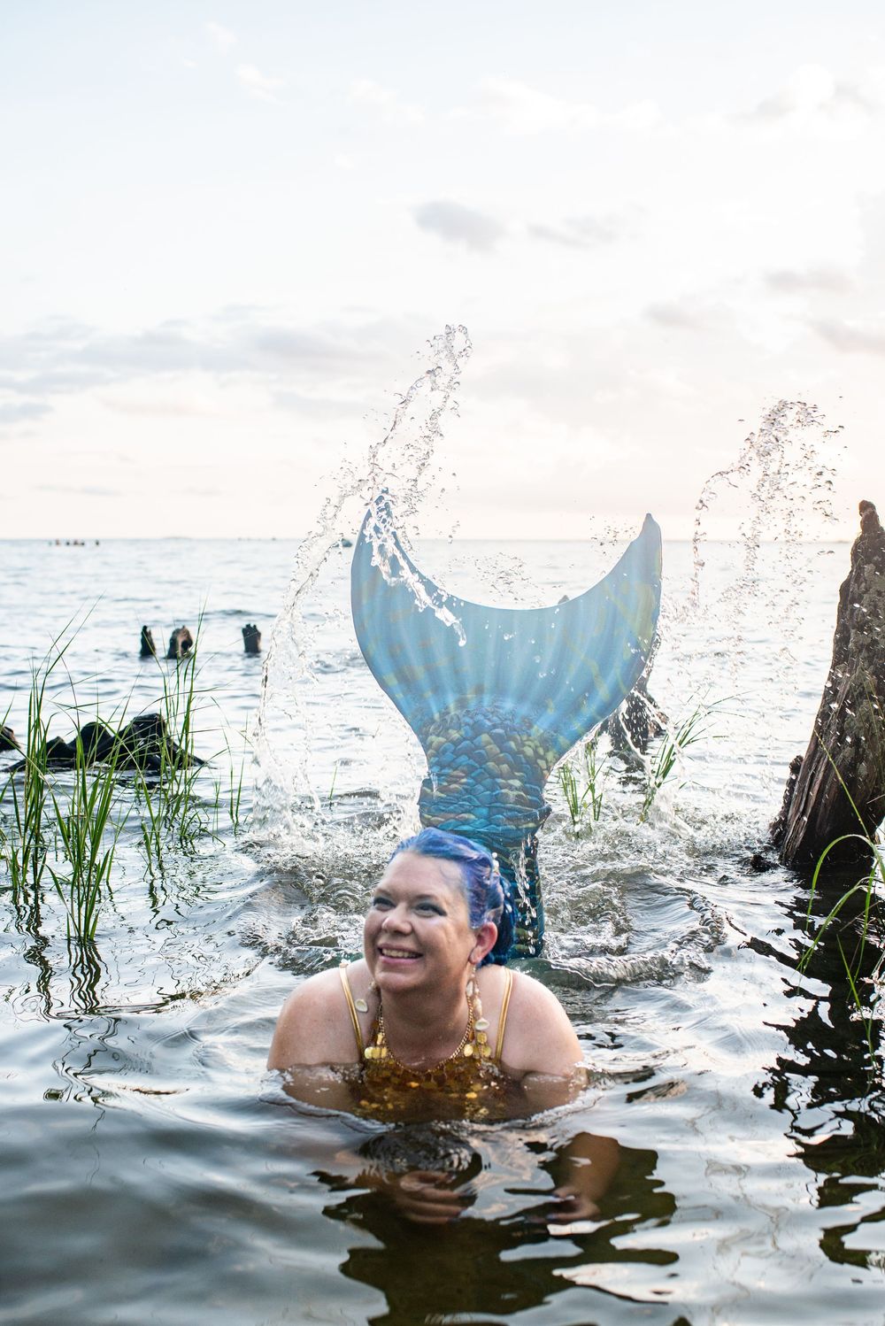 A person wearing a blue mermaid tail splashes in the water surrounded by reeds and ocean waves at sunset.