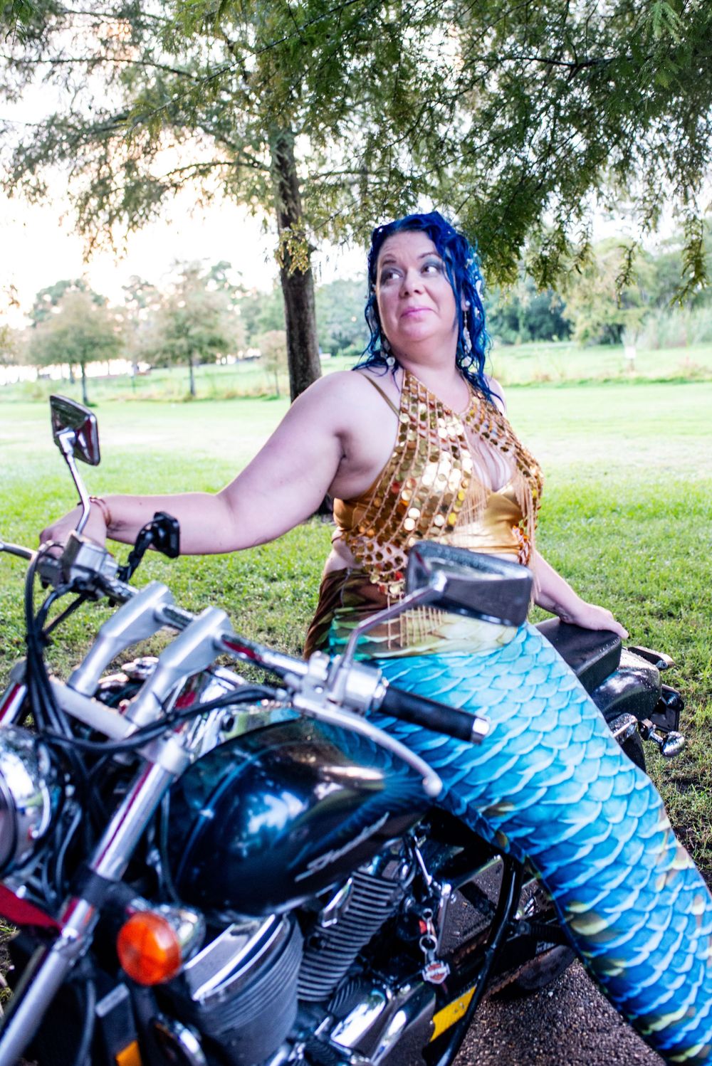 Person lounging on motorcycle in grassy field wearing blue patterned pants and gold top on sunny day.
