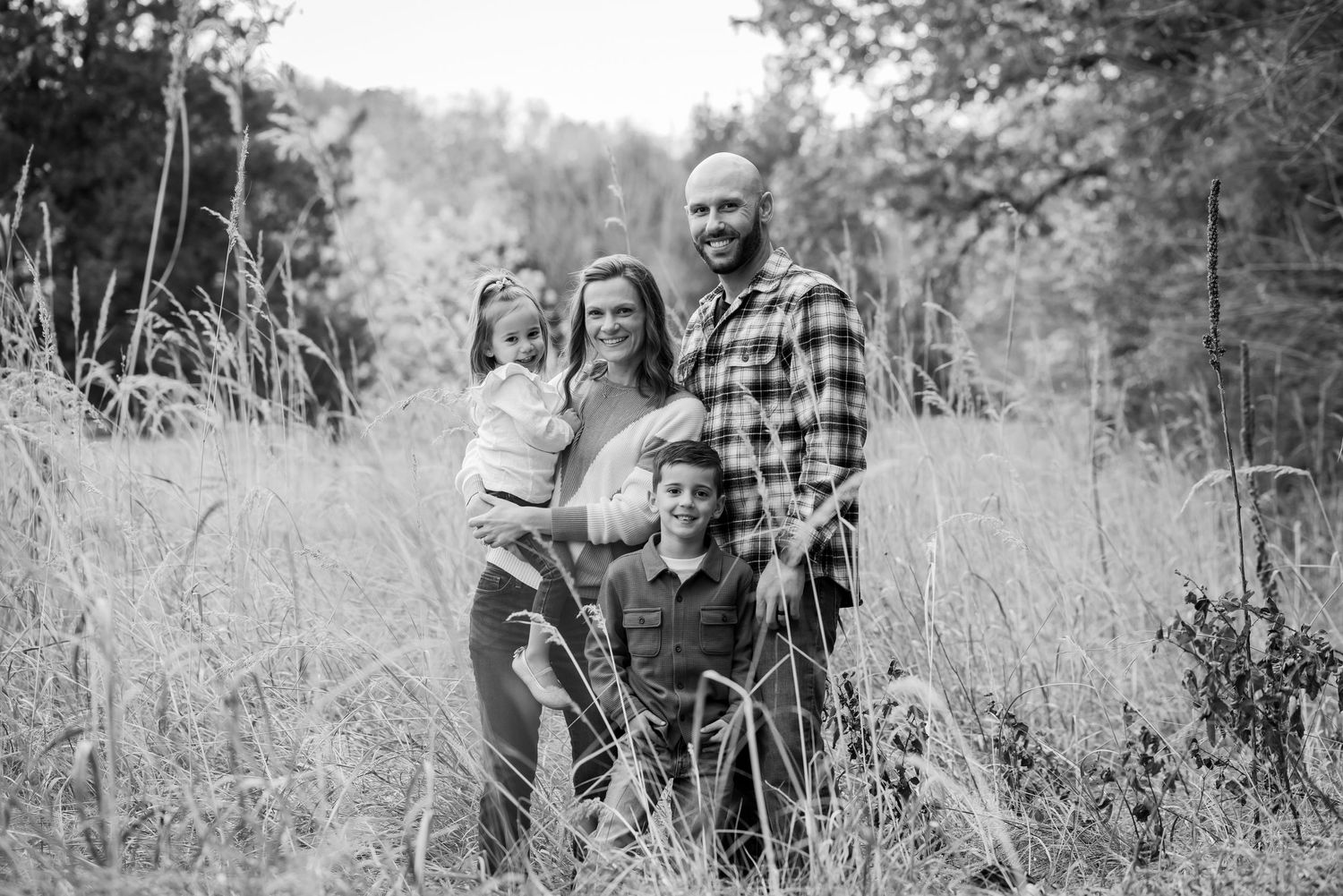 Black and white family portrait of five in tall grass field surrounded by trees.