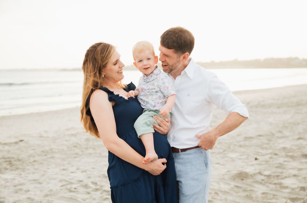 Pregnant woman in navy dress with husband and toddler son smiling together on a sandy beach. Second Beach Middletown, RI
