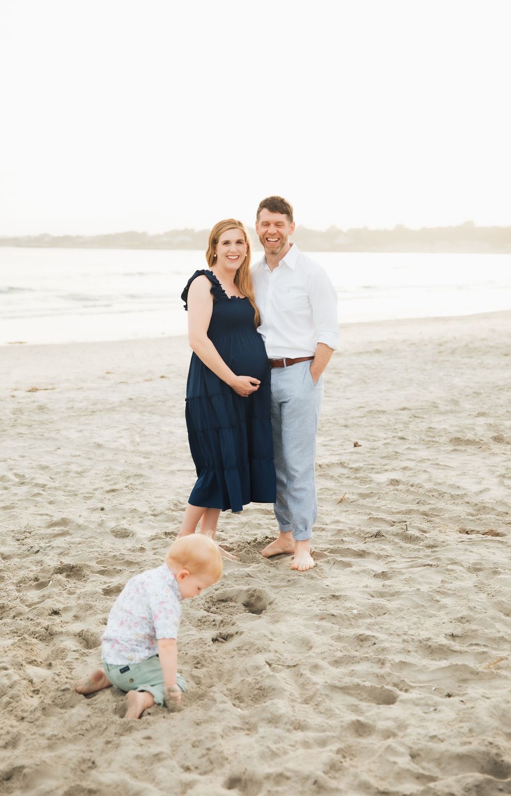 Pregnant couple smiling on a sandy beach while their toddler plays in the sand, casual family maternity photo session. Second Beach