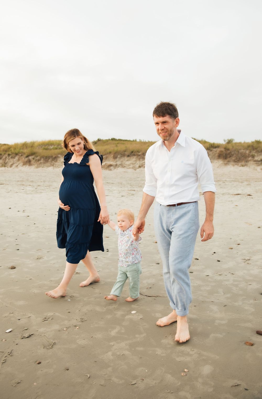 Pregnant woman in navy dress walks barefoot on beach with toddler and man in white shirt on cloudy day. Second Beach Middletown, RI