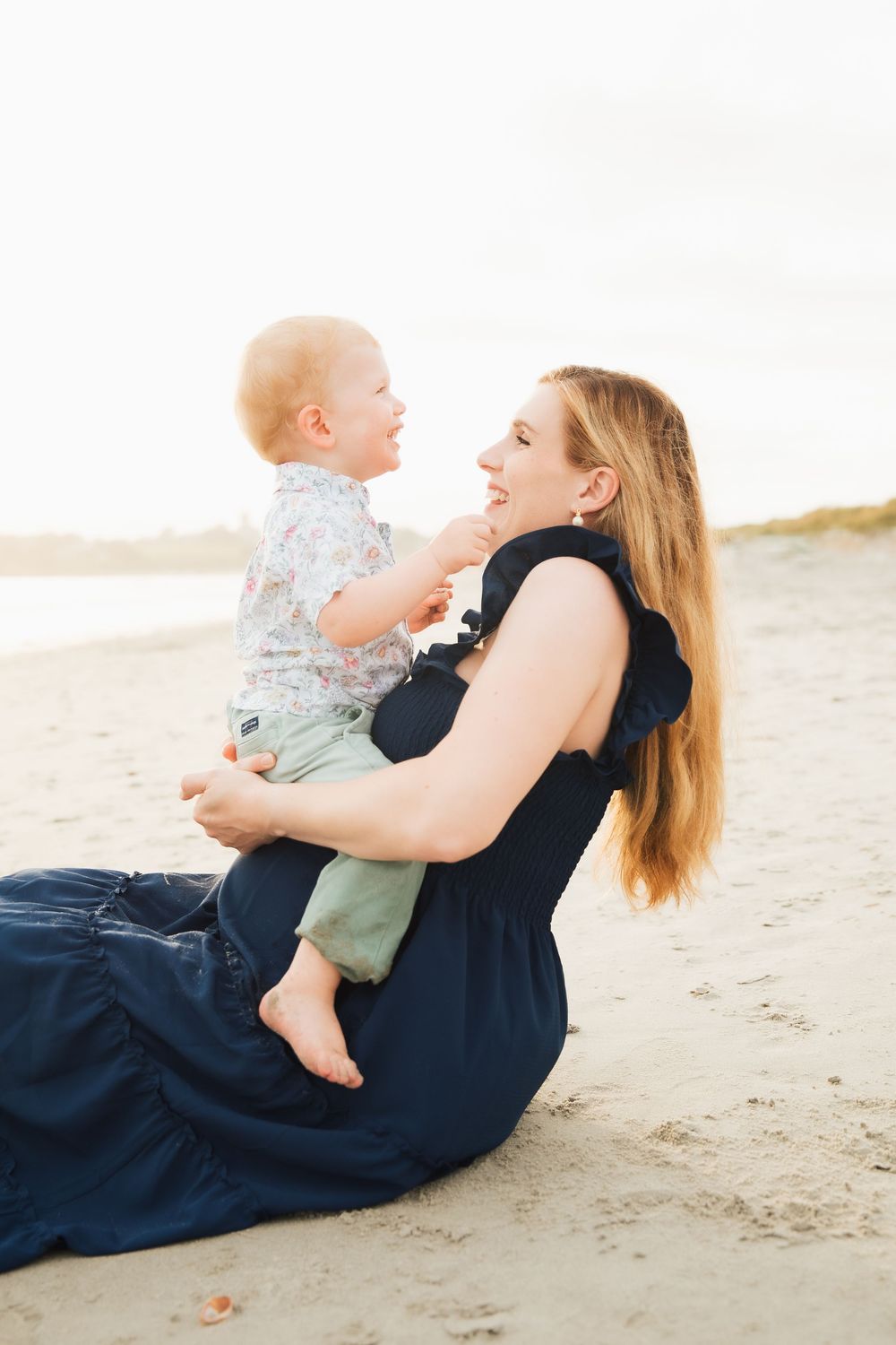 A mother sits on a sandy beach holding her smiling baby, both sharing a joyful moment in warm golden light Second Beach Middletown, RI.